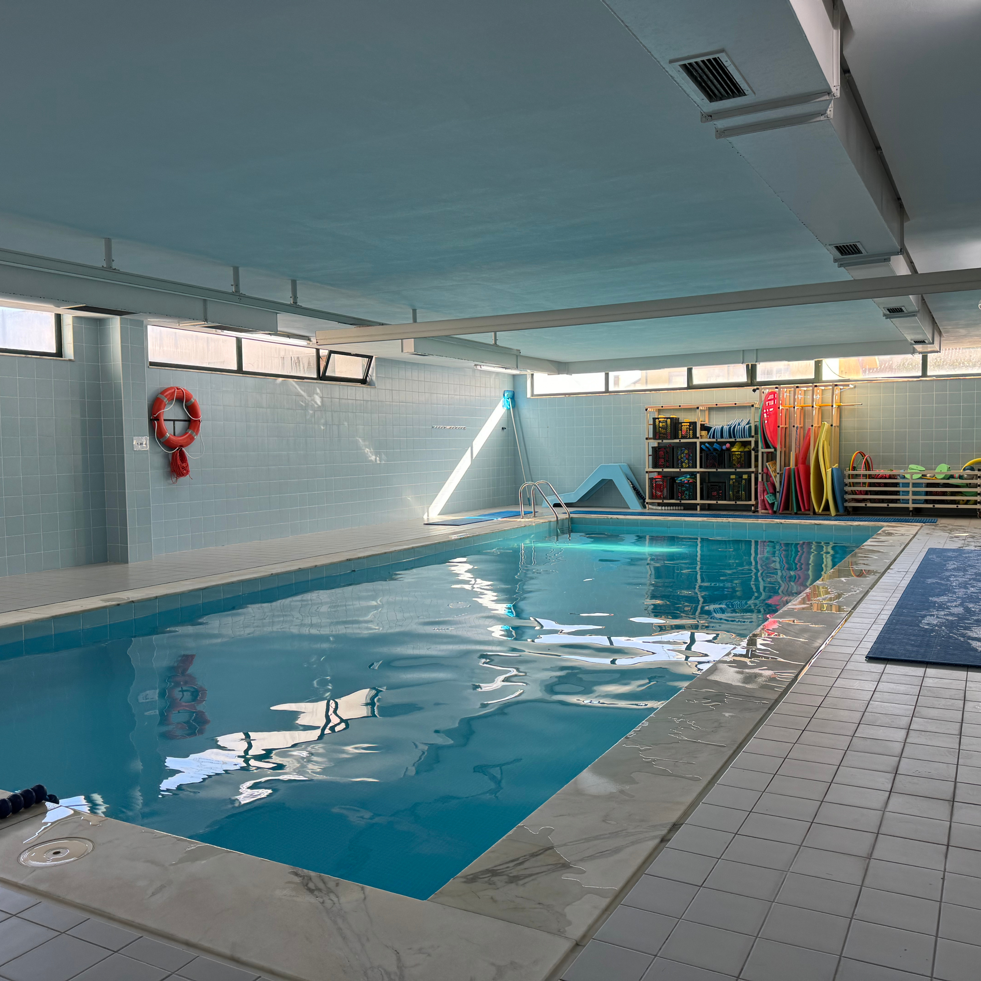 Fotografia de piscina interior. A piscina encontra-se com água mas sem utilizadores. Ao fundo, junto da parede, diverso material para práticas aquáticas.