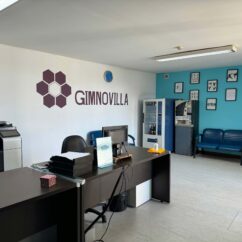 Espaço interior com duas mesas, uma cadeira, algum equipamento de apoio e um sofá de dois lugares. Na parede o logótipo e a palavra GimnoVilla.