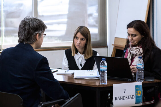 Três pessoas conversam junto de uma mesa. Na mesa estão pousados vários papéis. Uma das pessoas usa computador portátil.