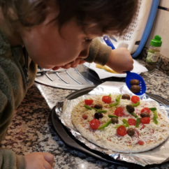 Criança segura na mão uma colher azul. Na referida colher estão duas azeitonas que estão a ser colocadas em pizza, ainda crua, pousada na mesa.