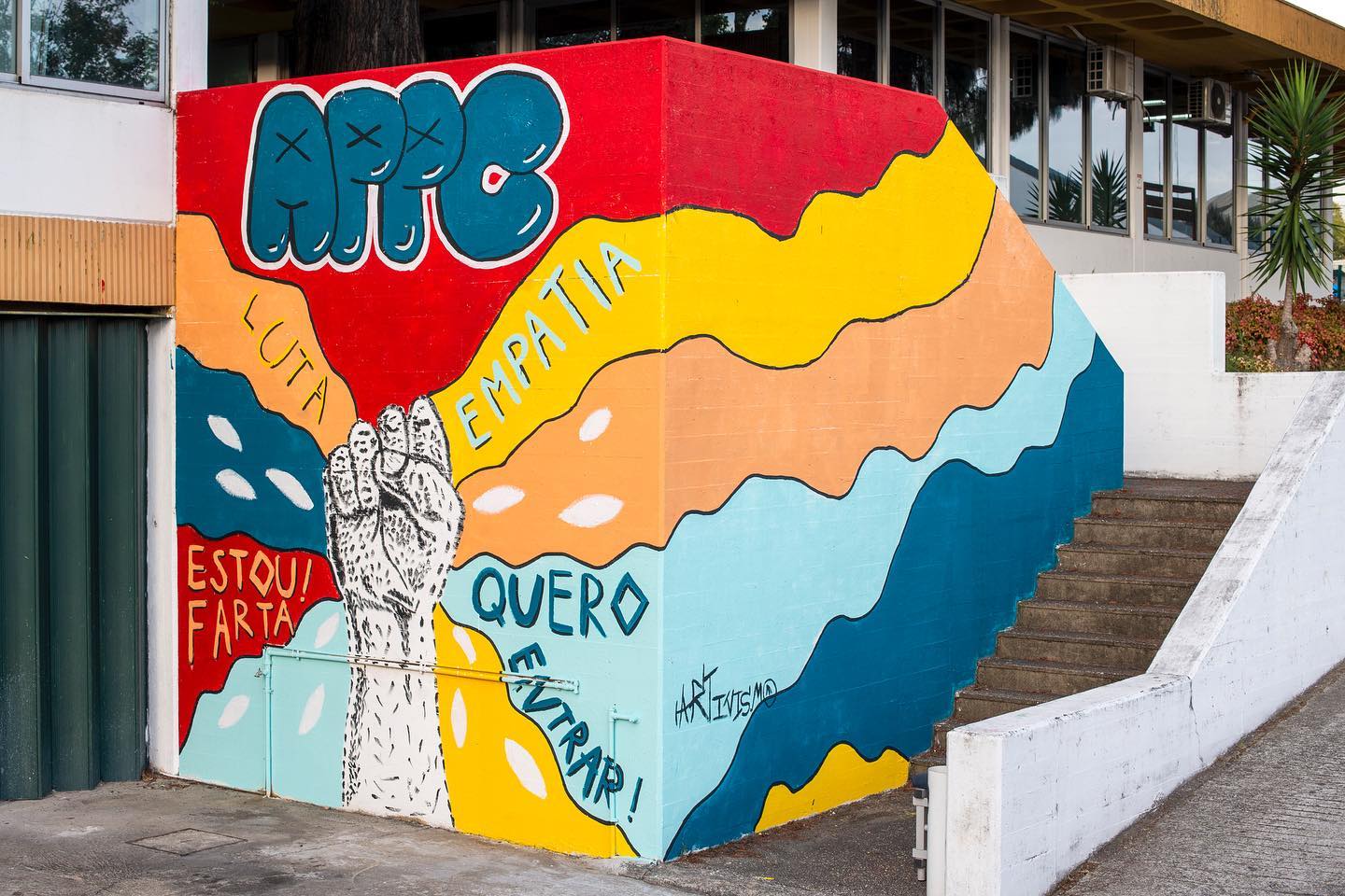 Muro colorido com pintura gigante. Escadas de acesso e alguns arbustos.