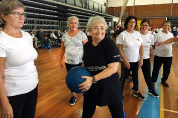 Seis pessoas em atividade física em pavilhão interior. Uma das pessoas segura uma bola azul. Ao fundo outras pessoas, sentadas e em pé, estão a assistir.