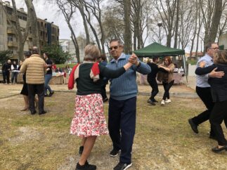 Várias pessoas dançam em jardim público. Árvores e edifícios ao fundo.