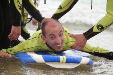 Pessoa deitada em prancha de surf a sorrir para a fotografia. Outras duas pessoas (parcialmente visíveis) prestam apoio.