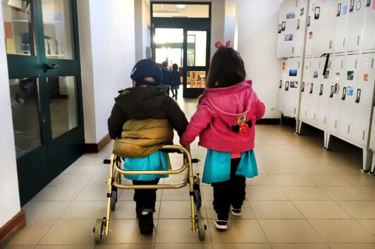 Duas crianças, de costas, caminham por um corredor. Uma das crianças usa andarilho. Cacifos à direita da imagem.