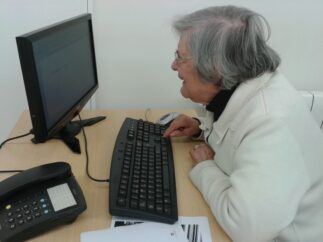 Pessoa sentada a olhar para ecrã de computador enquanto escreve no teclado. Na mesa vê-se ainda um telefone.