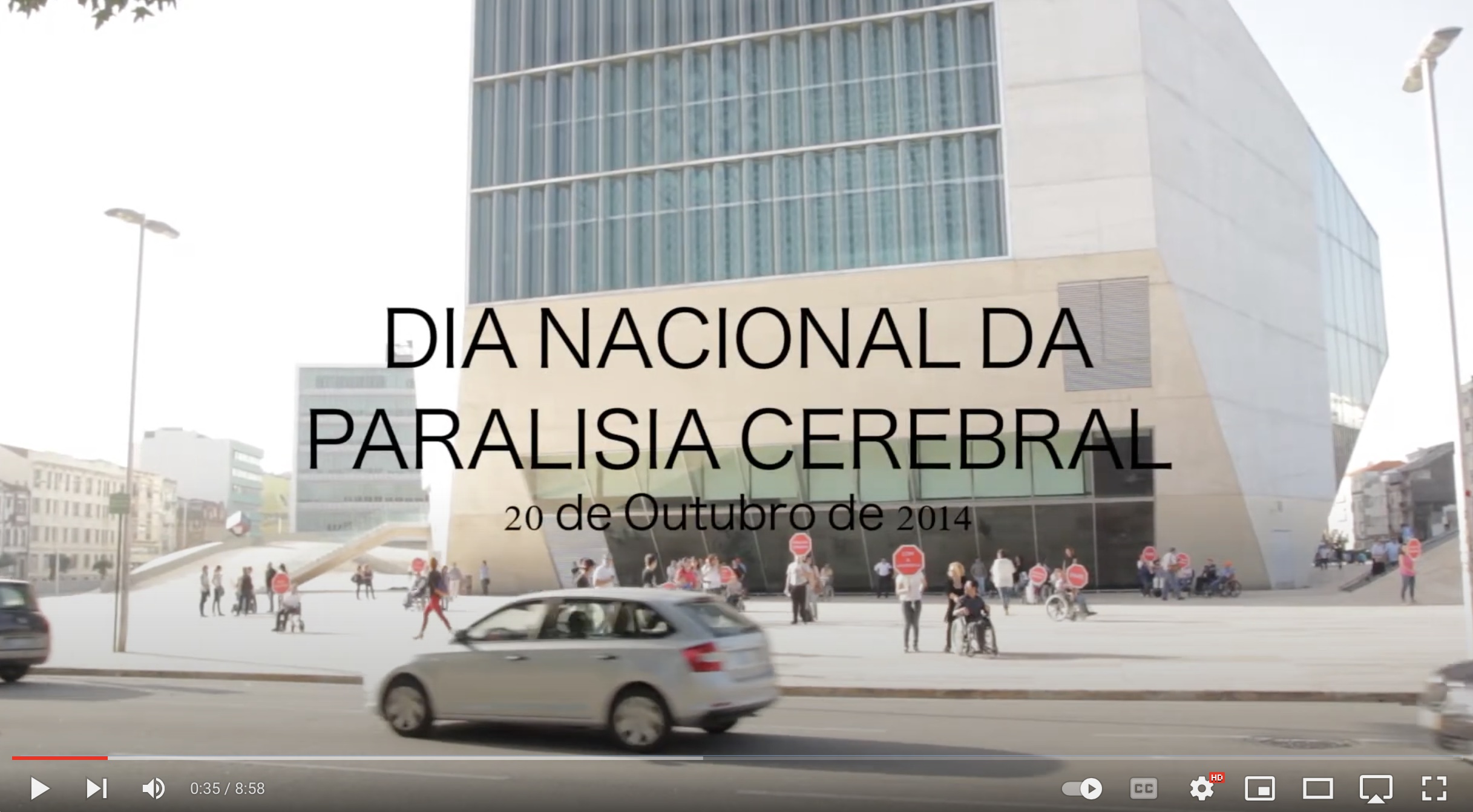 Captura de vídeo. Ao fundo um edifício (Casa da Música do Porto). À sua frente algumas pessoas seguram sinais de STOP com mensagens. Num plano mais próximo passa um carro. Ao centro da imagem, a preto, o título 