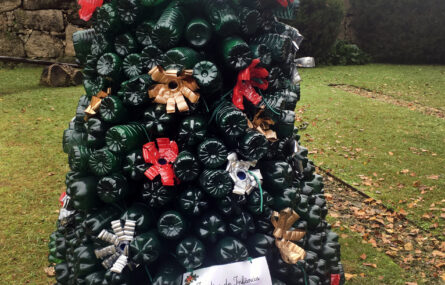 Uma suposta árvore feita integralmente com garrafas de plástico de tom verde escuro. No cimo tem uma estrela feita com tampas de garrafas pintadas em tom dourado. A árvore está colocada num jardim exterior. Ao fundo aparecem árvores e um muro.