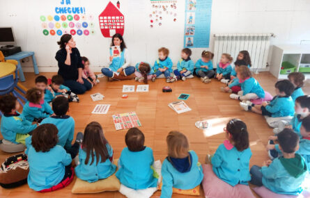Mais de duas dezenas de crianças sentadas em semicírculo no chão de uma sala. Surgem ainda duas pessoas, adultas, que aparentam fazer uma apresentação. Todos os rostos das crianças foram desfocados.