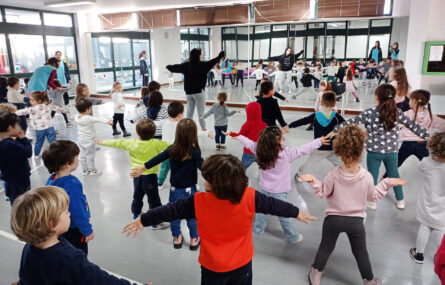 Perto de três dezenas de crianças a fazer giástica, ou dança, em espaço interior. Alguns adultos acompanham a atividade. A sala tem um espelho gogante a todo o comprimento da parede do fundo – sendo que a imagem do grupo é refletida no referido espelho. Todos os rostos das crianças foram desfocados.