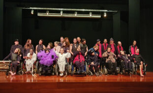 Perto de três dezenas de pessoas, em palco, alinhadas para a fotografia. Essencialmente são atores e atrizes da Companhia Era uma vez Teatro da APPC. Aparecem, igualmente, outros elementos da equipa técnica e do grupo musical que apoiou a apresentação. Ao fundo a imagem alguns holofotes.