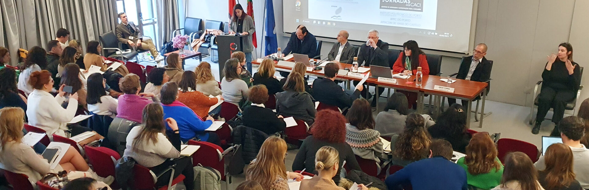 Fotografia de auditório interior. Várias pessoas sentadas nas cadeiras do auditório. Ao fundo, na zona de palco, mesa com cinco intervenientes sentados. De pé, junto de um púlpito, está uma pessoa a falar. Do lado direito da imagem, sentada a gesticular, está intérprete de Língua Gestual Portuguesa.