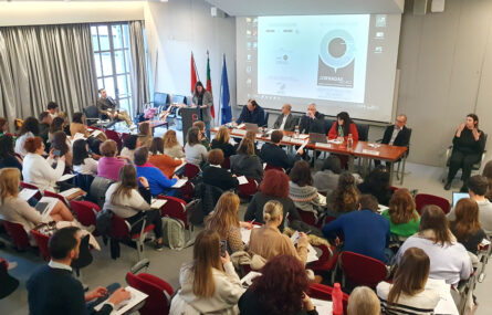 Fotografia de auditório interior. Várias pessoas sentadas nas cadeiras do auditório. Ao fundo, na zona de palco, mesa com cinco intervenientes sentados. De pé, junto de um púlpito, está uma pessoa a falar. Do lado direito da imagem, sentada a gesticular, está intérprete de Língua Gestual Portuguesa.