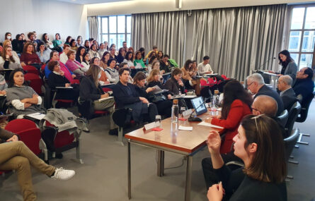 Fotografia de auditório interior. Várias pessoas sentadas nas cadeiras do auditório. Na zona de palco, de costas, mesa com cinco intervenientes sentados. De pé, junto de um púlpito, está uma pessoa a falar. Do lado esquerdo da imagem, em primeiro plano, vê-se intérprete de Língua Gestual Portuguesa a gesticular.