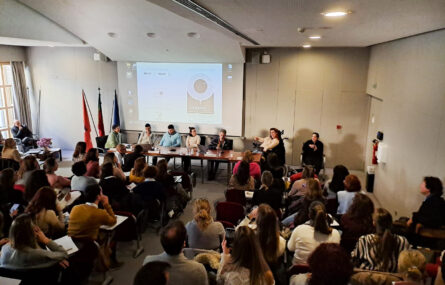 Fotografia de auditório interior. Várias pessoas sentadas nas cadeiras do auditório. Ao fundo, na zona de palco, mesa com cinco intervenientes sentados e uma outra pessoa a usar cadeira de rodas. Do lado direito da imagem, sentada a gesticular, está intérprete de Língua Gestual Portuguesa.