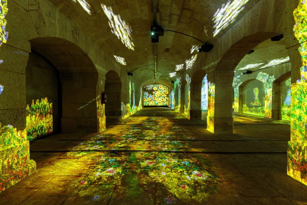 Vista de galeria interior com imagens projetadas no chão, paredes laterais, arcadas e tetos. Tons marcadamente amarelos e verdes em ambiente de notória escuridão.