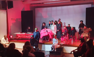 Fotografia de 19 atores e atrizes em palco, já na fase final da peça. À direita conseguem-se ainda ver alguns dos elementos da banda que atuou ao vivo.