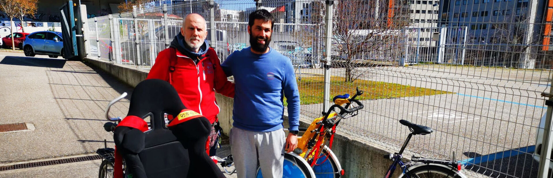 Duas pessoas, em espaço exterior, junto de várias bicicletas adaptadas. Mais ao fundo da imagem são visíveis alguns carros.