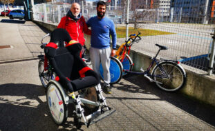 Duas pessoas, em espaço exterior, junto de várias bicicletas adaptadas. Mais ao fundo da imagem são visíveis alguns carros.