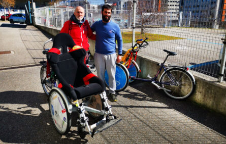 Duas pessoas, em espaço exterior, junto de várias bicicletas adaptadas. Mais ao fundo da imagem são visíveis alguns carros.