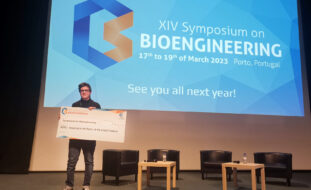 Fotografia de pessoa em pé, num palco, a segurar exemplar (tamanho grande) de um cheque referente ao donativo oferecido à APPC. Ao fundo vê-se projeção do cartaz do XIV Symposium on Bioengineering.