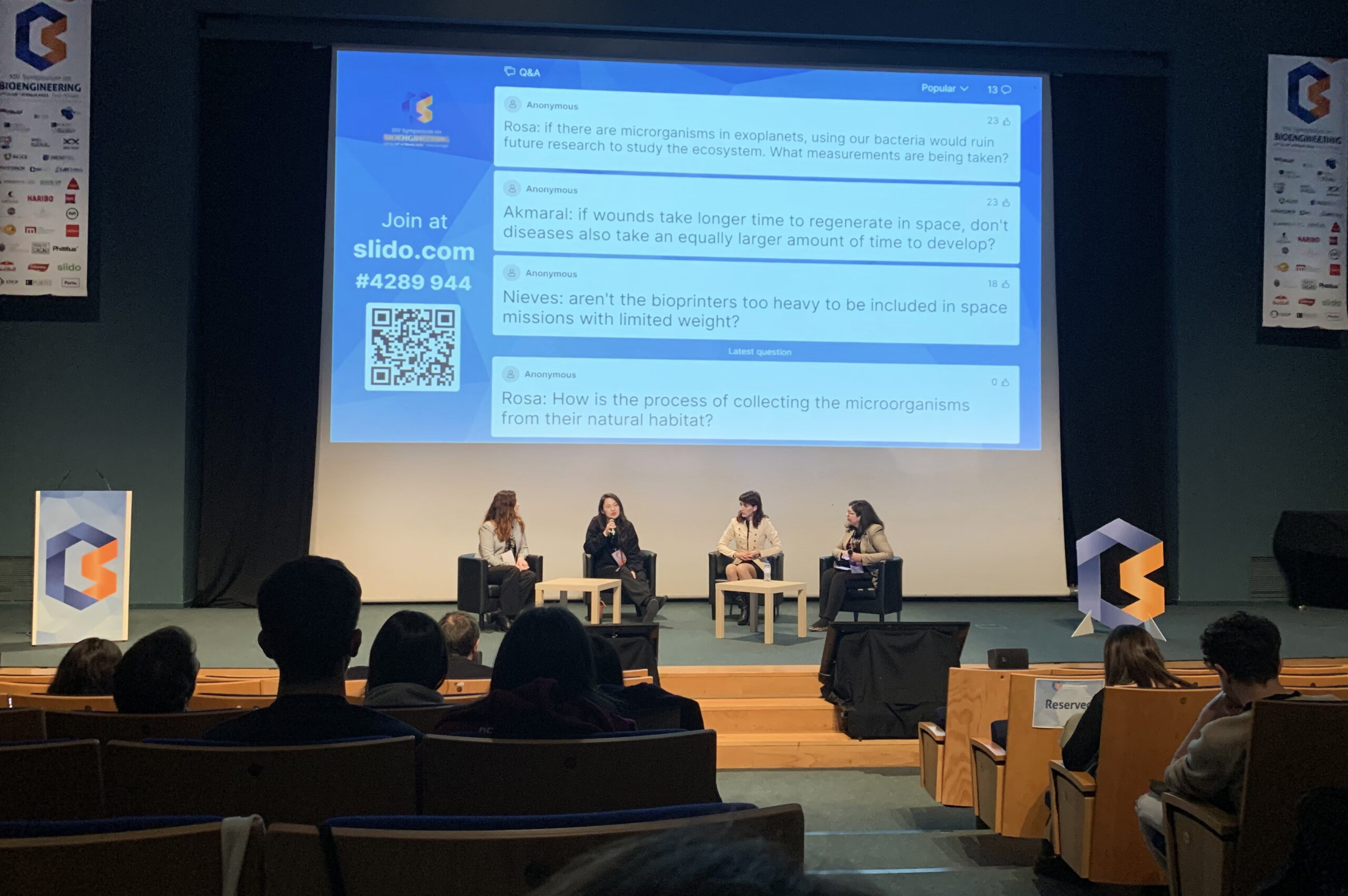 Em primeiro plano, pessoas sentadas de costas. Ao fundo, em palco, quatro pessoas sentadas a conversar e projeção do assunto em debate.