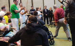 Foto vertical de palco (em auditório, interior) de pessoas a tocar e a praticar Capoeira. Ao fundo, junto à parede lateral, outras pessoas sorriem e aplaudem.