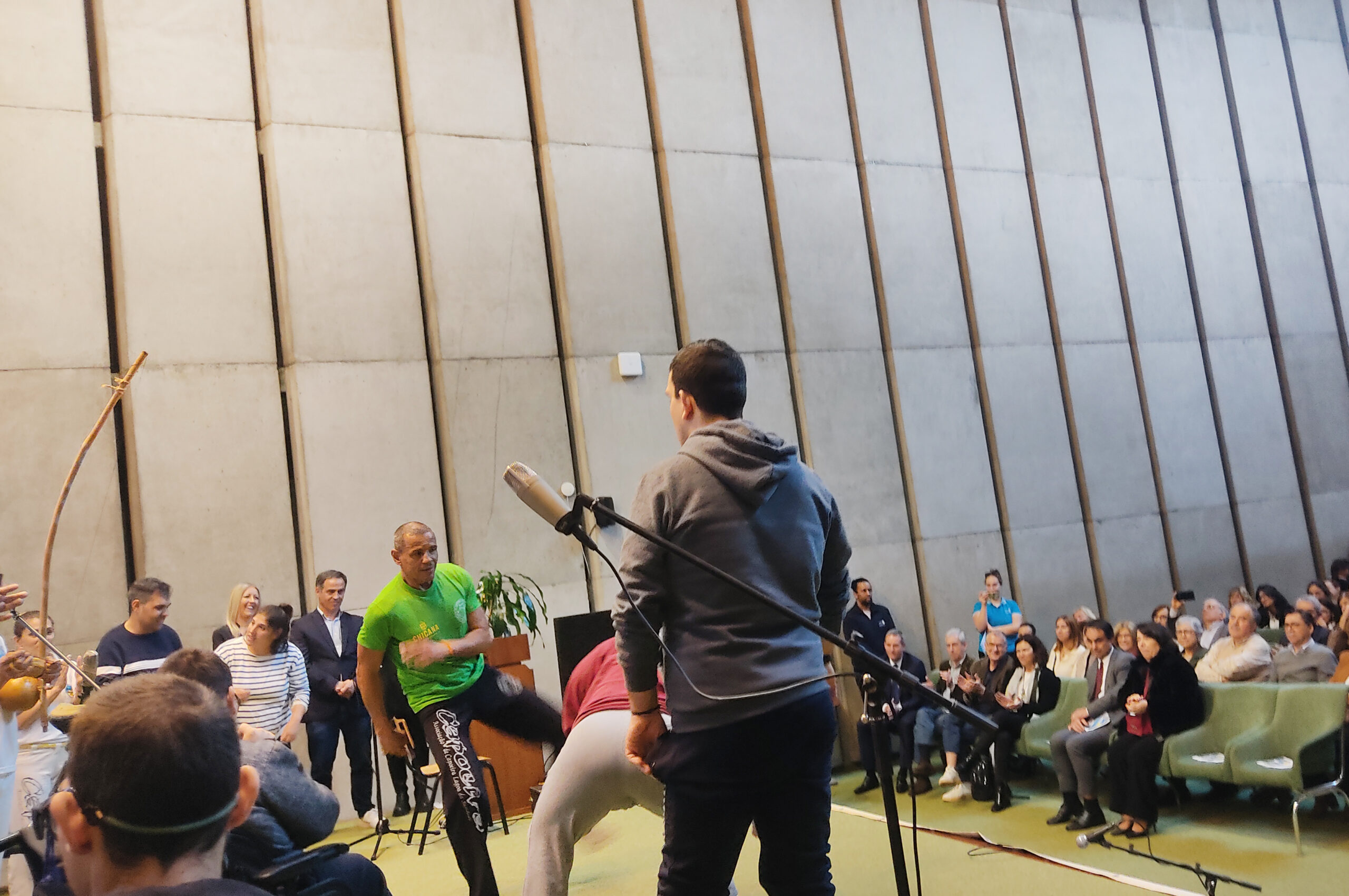 Foto de palco (em espaço interior) de pessoas a tocar e a praticar Capoeira. Ao fundo, sentadas nas primeiras filas de cadeiras do auditório, várias pessoas assistem e aplaudem.