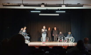 Fotografia de palco, com fundo negro, com atores e atrizes igualmente vestidos de negro a fazerem um semi-círculo. Ao centro, em destaque, uma atriz faz a leitura da mensagem do Dia Mundial do Teatro 2023.
