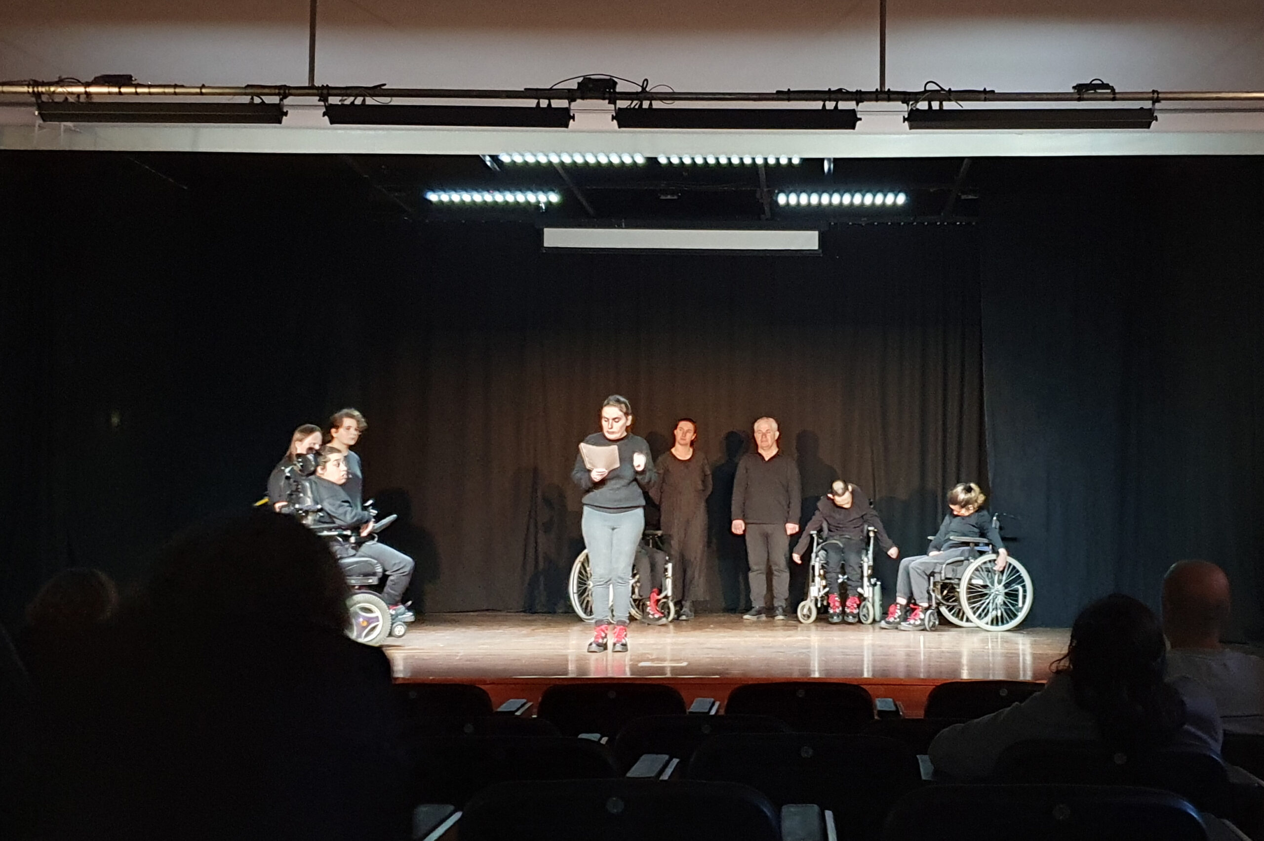 Fotografia de palco, com fundo negro, com atores e atrizes igualmente vestidos de negro a fazerem um semi-círculo. Ao centro, em destaque, uma atriz faz a leitura da mensagem do Dia Mundial do Teatro 2023.