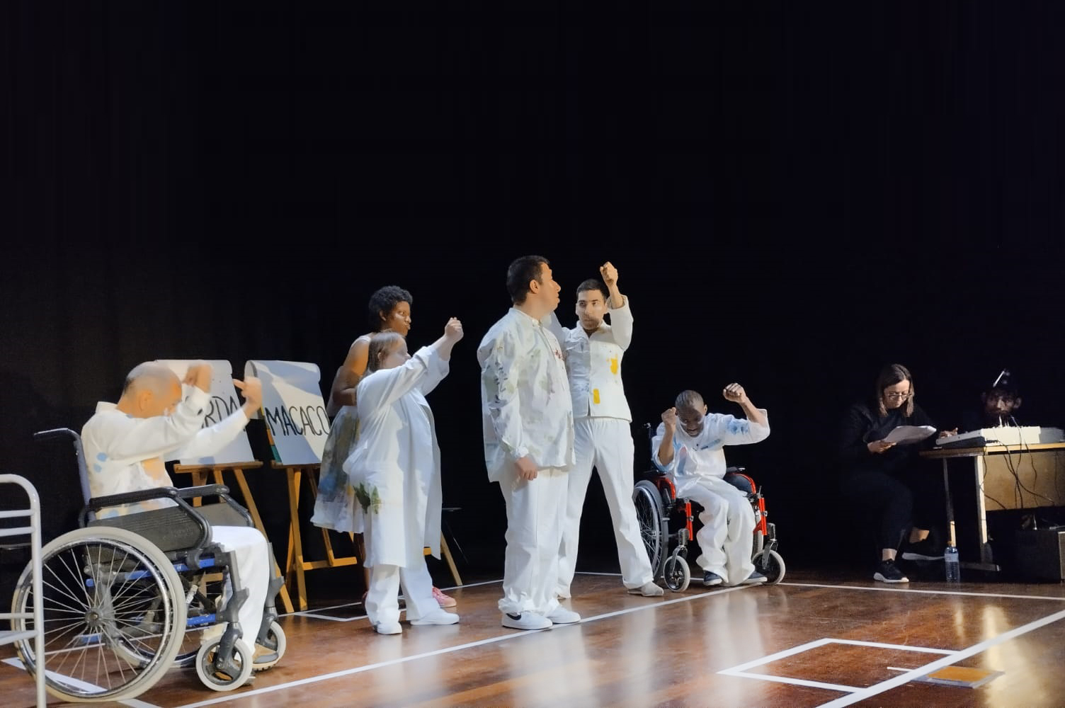 Palco com atores e atrizes, vestidos de branco, em pleno desempenho. Ao fundo, à direita, estão dois elementos da equipa de apoio à peça.