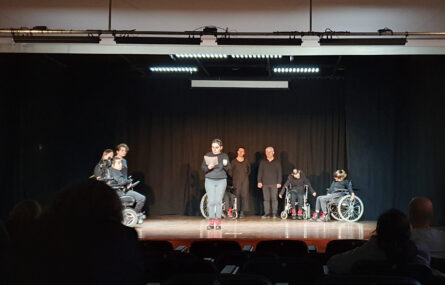 Atriz e Fotografia de palco, com fundo negro, com atores e atrizes (igualmente vestidos de negro) a comporem um semi-círculo. Ao centro, em destaque, uma atriz faz a leitura da mensagem do Dia Mundial do Teatro 2023.