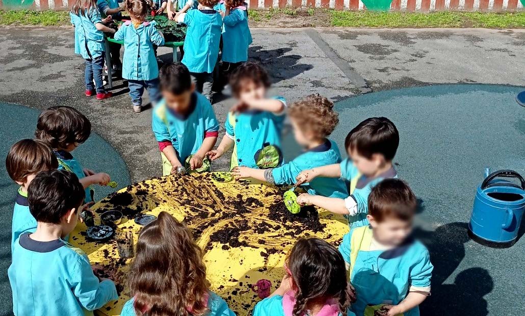 Fotografia de dois grupos de crianças, em redor de duas mesas (a do fundo, verde; a mais próxima, amarela), a brincarem com terra. Todos os rostos das crianças aparecem desfocados.