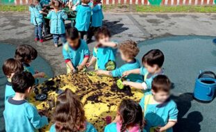 Fotografia de dois grupos de crianças, em redor de duas mesas (a do fundo, verde; a mais próxima, amarela), a brincarem com terra. Todos os rostos das crianças aparecem desfocados.