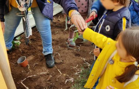 Uma senhora coloca na mão de uma criança um pouco de terra. Mais ao fundo outras crianças brincam também com a terra do jardim. Todos os rostos das crianças aparecem desfocados.