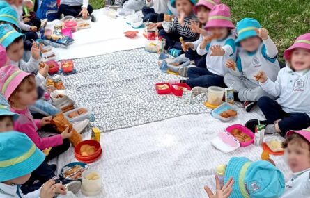 Dois grupos de crianças sentadas na relva com o lanche pousado em toalhas gigantes. Algumas das crianças dizem “adeus” para a fotografia. Todos os rostos das crianças aparecem desfocados.