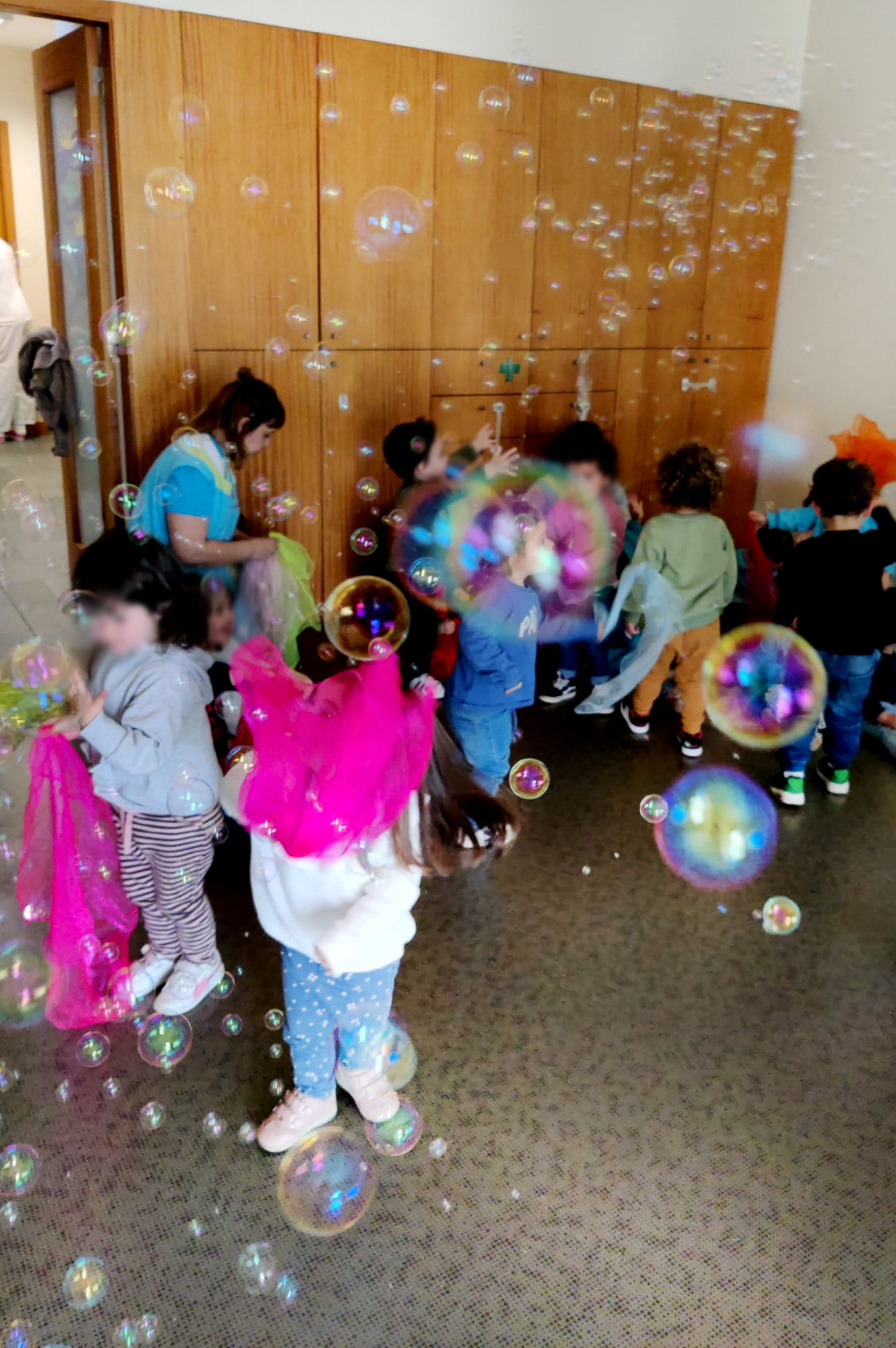 Em espaço interior um grupo com várias crianças diverte-se a perseguir bolas de sabão. Todos os rostos das crianças aparecem desfocados.