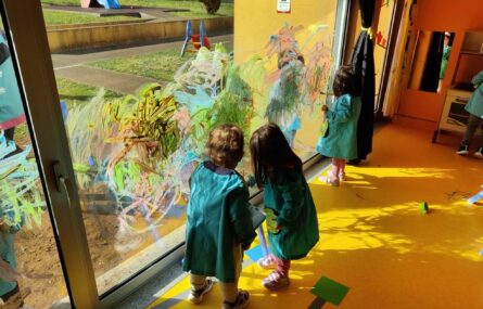 Três crianças (de costas) fazem pinturas e desenhos no vidro que separa um espaço interior do jardim.