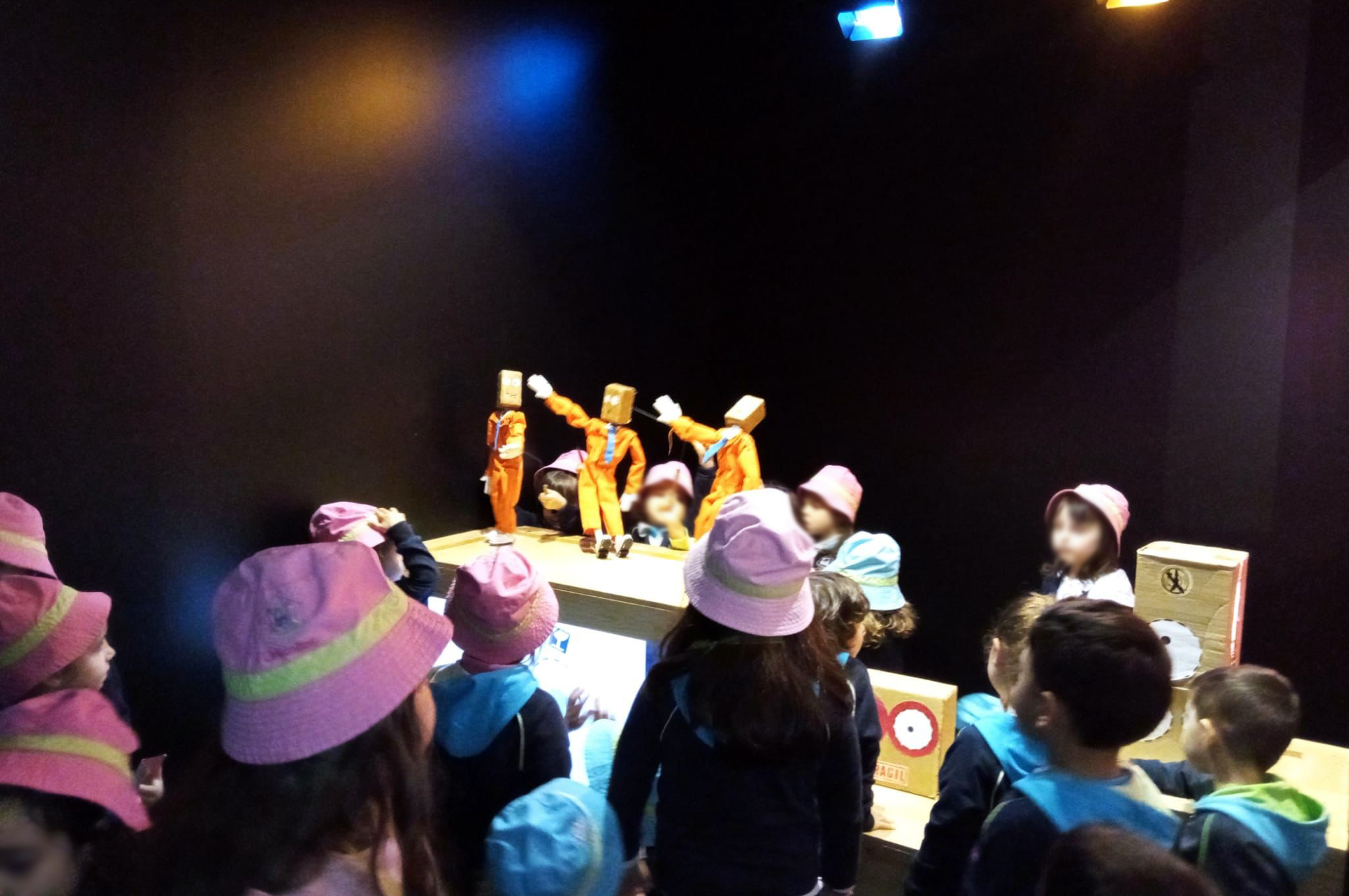 Em espaço interior, escuro, grupo de crianças aprecia marionetas e alguns adereços. Todos os rostos das crianças aparecem desfocados.