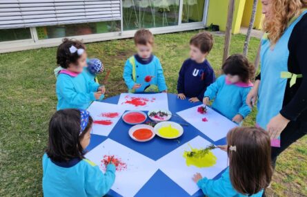 Grupo de crianças a fazer pinturas em papel. A mesa que usam está colocada no relvado de um jardim. Uma adulta acompanha-os. Todos os rostos das crianças aparecem desfocados