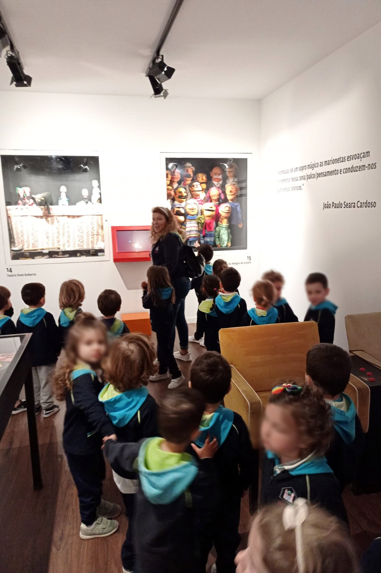 Fotografia de crianças em visita a exposição (em espaço interior). Na parede do fundo encontram-se dois cartazes gigantes e na lateral uma mensagem (parcialmente ilegível). Todos os rostos das crianças aparecem desfocados.
