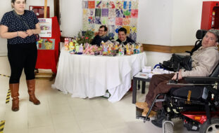 Fotografia da “banquinha” de venda de produtos do Centro de Atividades e Capacitação para a Inclusão (CACI) Delfim Maia da Associação do Porto de Paralisia Cerebral. À esquerda, de pé, uma jovem a sorrir para a fotografia. Sentados, na mesa de venda de produtos, dois sorridentes clientes do CACI. À direita, outro cliente da APPC