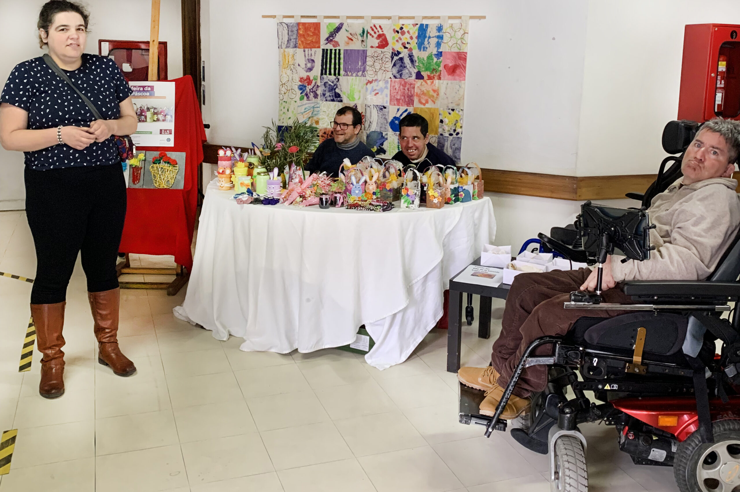 Fotografia da “banquinha” de venda de produtos do Centro de Atividades e Capacitação para a Inclusão (CACI) Delfim Maia da Associação do Porto de Paralisia Cerebral. À esquerda, de pé, uma jovem a sorrir para a fotografia. Sentados, na mesa de venda de produtos, dois sorridentes clientes do CACI. À direita, outro cliente da APPC