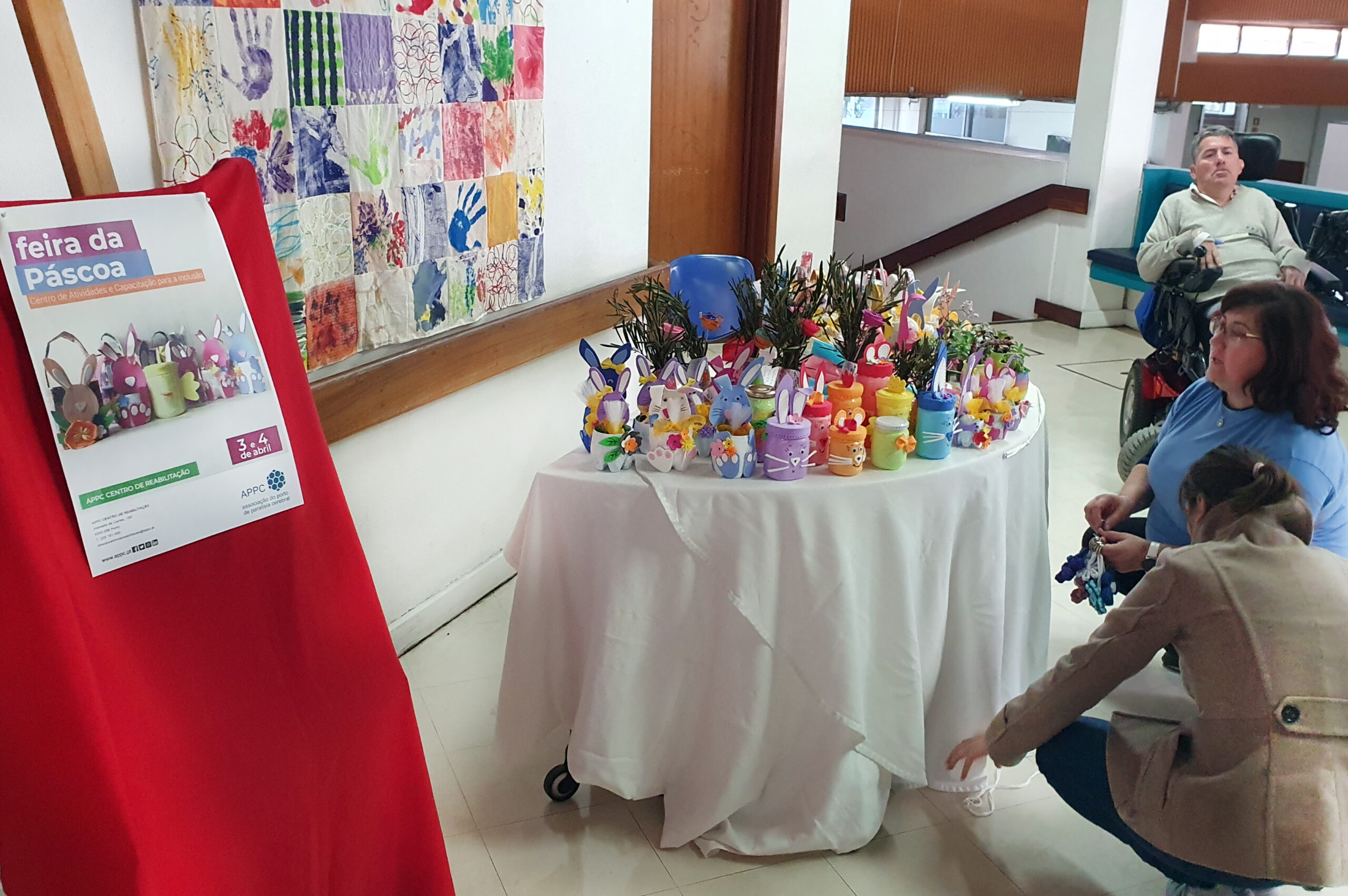 Preparativos finais da mesa com produtos para venda na “Feira da Páscoa”. À esquerda, suporte com cartaz da atividade. Mais à direita, duas pessoas fazem os arranjos finais na apresentação da mesa de produtos a vender.