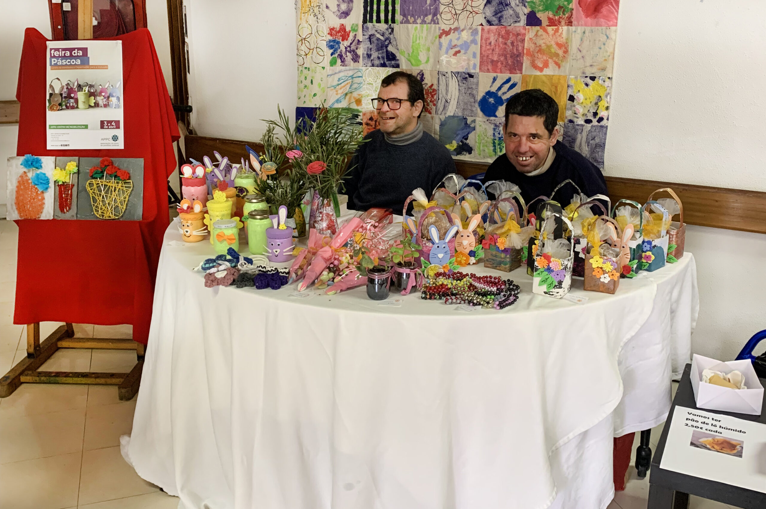 Sentados atrás da mesa com produtos para venda estão dois clientes da APPC. Em cima da mesa estão inúmeros produtos relacionados com a Páscoa.