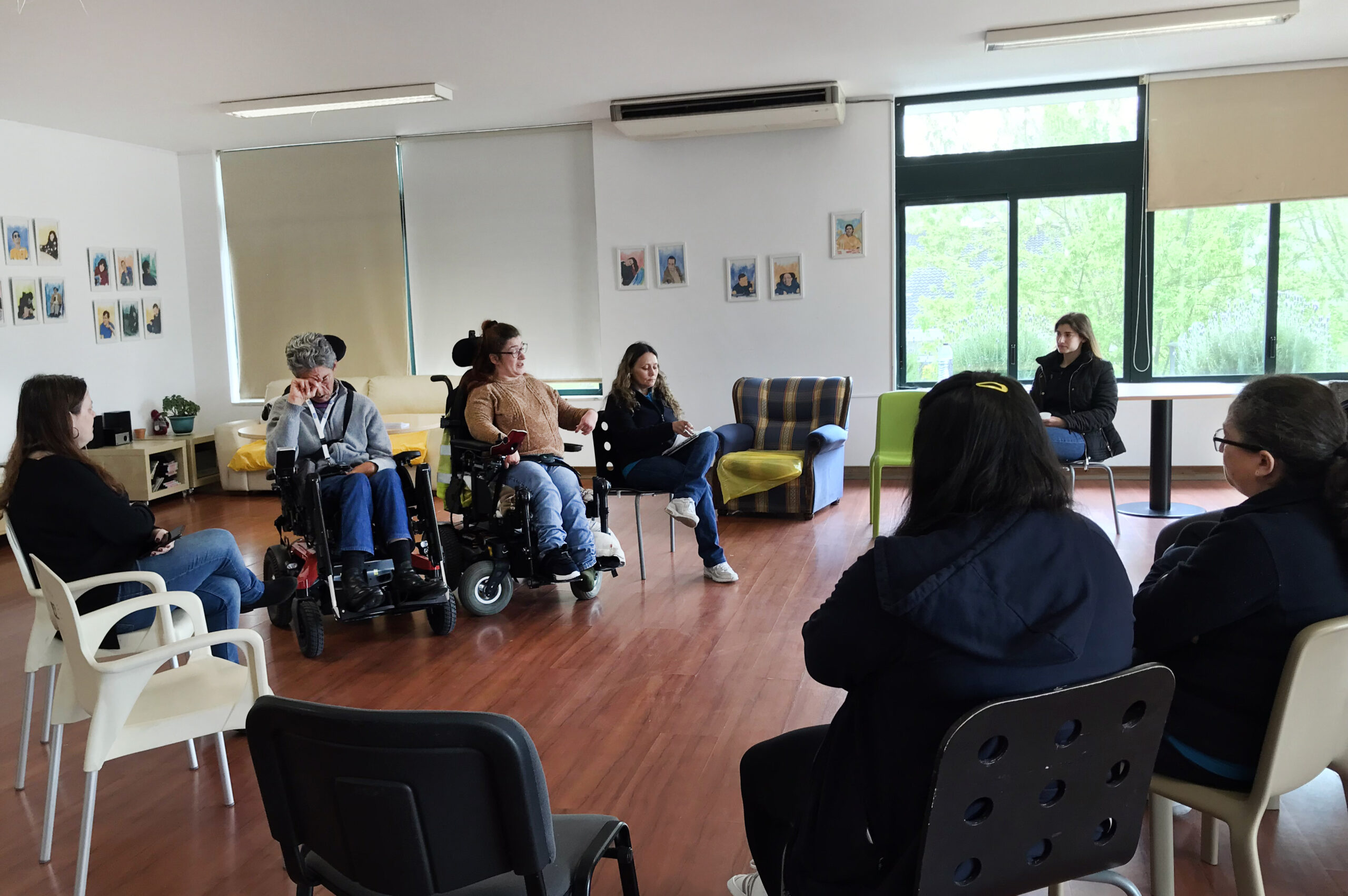 Sete pessoas sentadas, em círculo, em espaço interior da Villa Urbana. Algumas cadeiras vazias. Ao fundo, na parede, inúmeros retratos estilizados de clientes da unidade residencial da APPC.