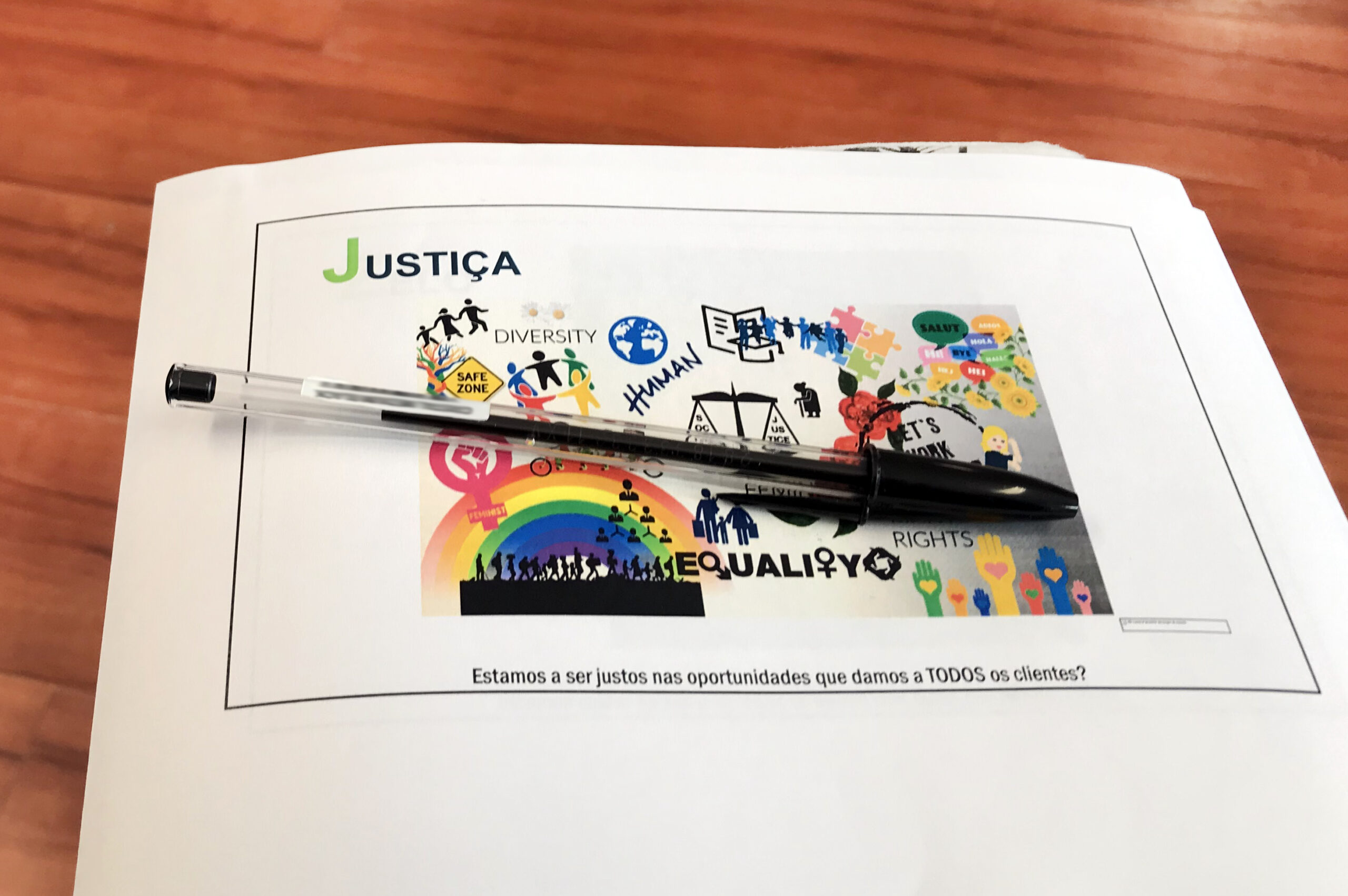 Pousado em cima de uma mesa está um documento com imagens sobre justiça, igualdade e diversidade. Vê-se, ainda, uma esferográfica.