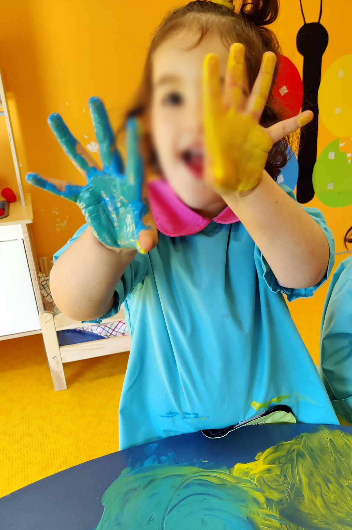 Grande plano de criança a mostrar as mãos pintadas com tinta azul e amarela. Rosto desfocado.
