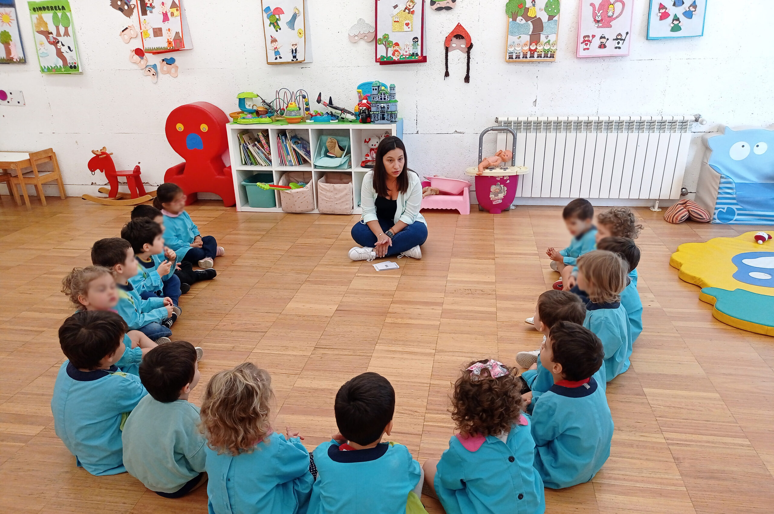 Crianças sentadas em semi-círculo no chão a ouvir uma pessoa adulta a falar. Fotografia de interior com parede repleta de trabalhos e pinturas. Rostos das crianças desfocados.