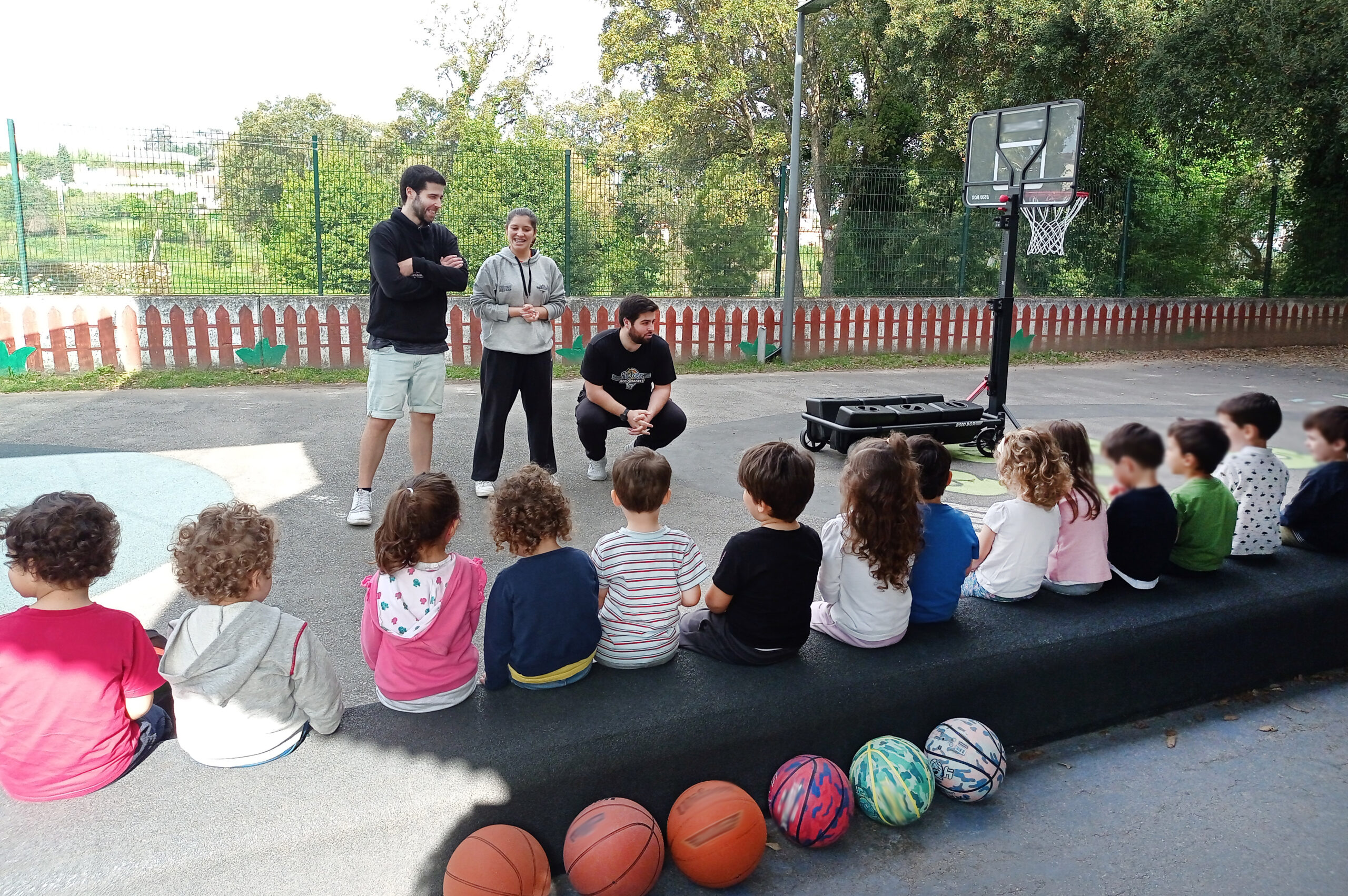 Várias crianças sentadas de costas. Três adultos falam com elas. Ao fundo, do lado direito, tabela portátil de basquetebol. Em primeiro plano estão pousadas seis bolas, coloridas, de basquetebol. Rostos das crianças desfocados.